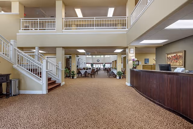 Spacious interior of a senior living facility with a patterned carpeted floor, a wooden reception desk on the right, a staircase with white railings on the left, and a dining area with tables and chairs visible in the background under a high ceiling with fluorescent lighting.