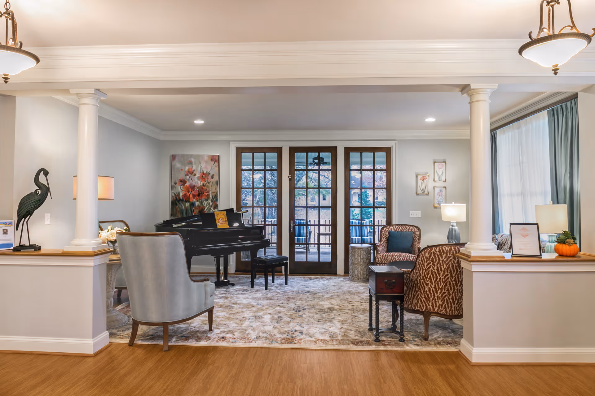 A cozy living room area in Larmax Homes featuring a grand piano, several upholstered chairs, a patterned rug, and large glass doors leading to an outdoor patio. The room is decorated with artwork, lamps, and a small pumpkin on a side table, with soft lighting from ceiling fixtures and table lamps.