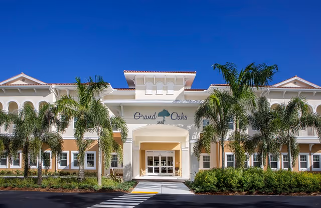 Front exterior view of the Grand Oaks senior living facility building with palm trees and landscaping under a clear blue sky.