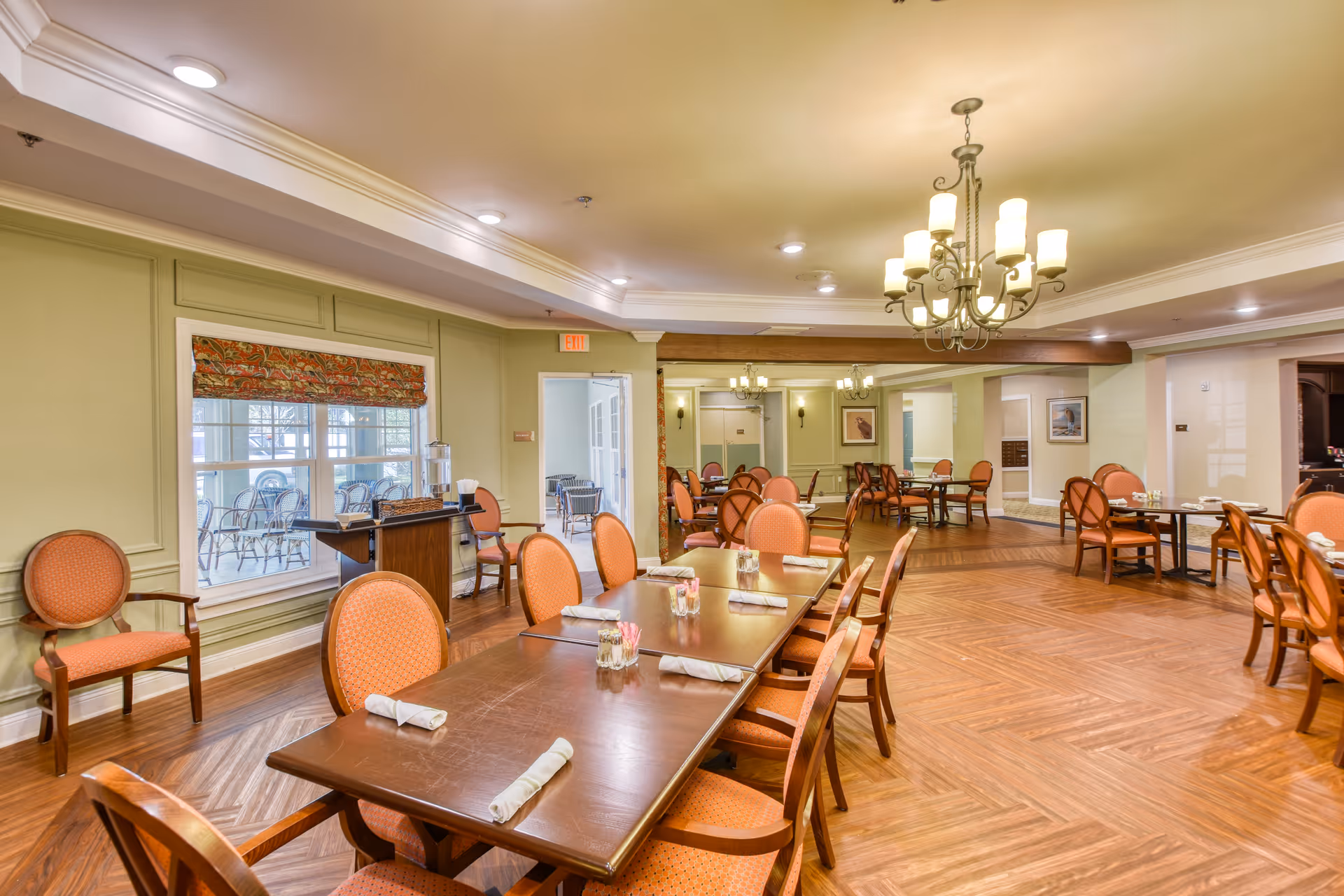 A spacious dining room with multiple wooden tables and orange cushioned chairs arranged neatly. The room features hardwood flooring, soft yellow walls, and a chandelier hanging from the ceiling. Large windows with patterned valances allow natural light to enter, and there is a water dispenser on a small table near the window.