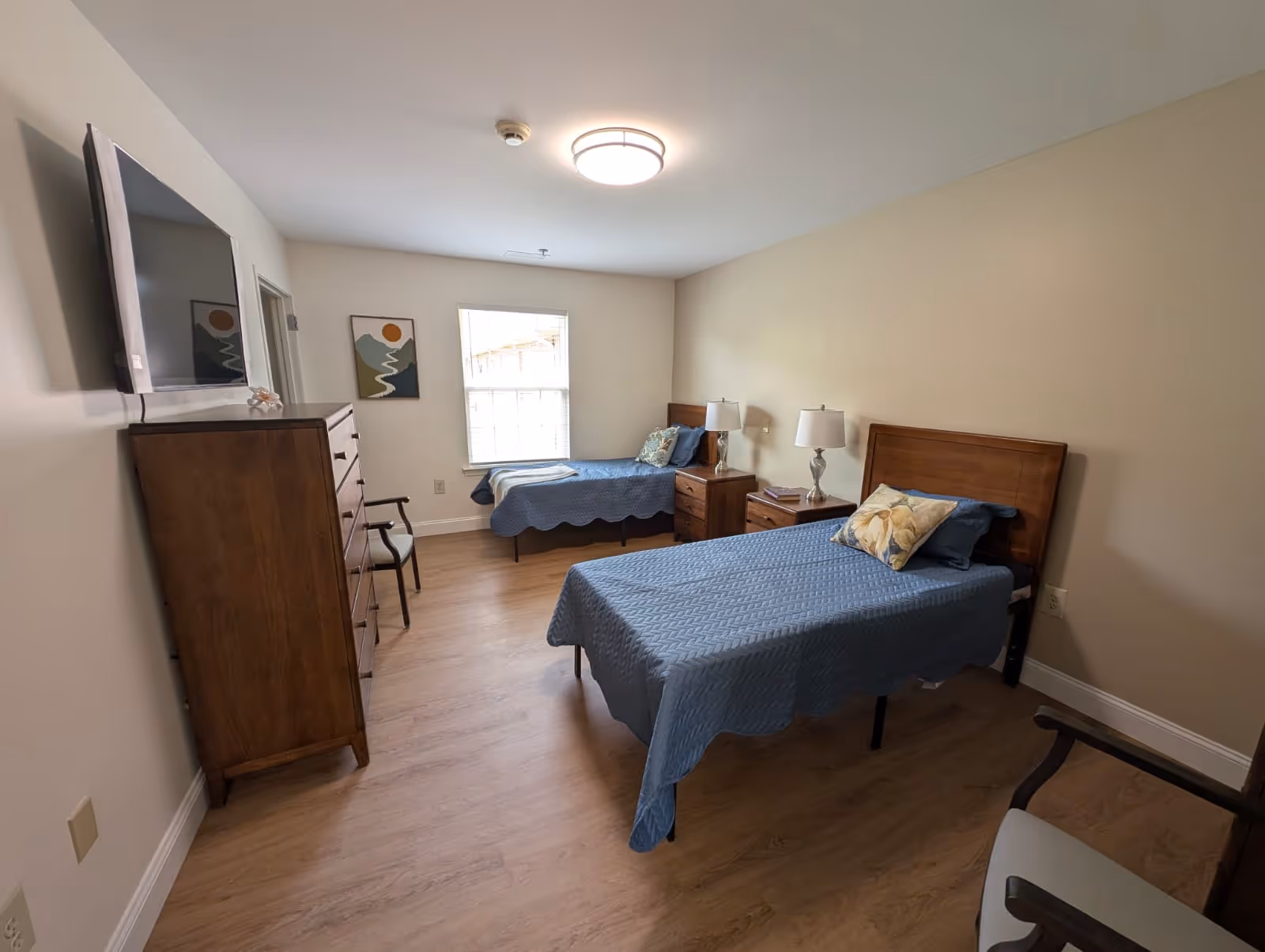 A bedroom with two single beds covered in blue quilts, each with a pillow. Between the beds are two wooden nightstands with lamps. A wooden dresser with a flat-screen TV mounted above it is on the left wall. There is a window with blinds on the far wall and a framed abstract landscape painting next to it. The floor is wood, and there are two chairs visible, one near the dresser and one in the foreground on the right.
