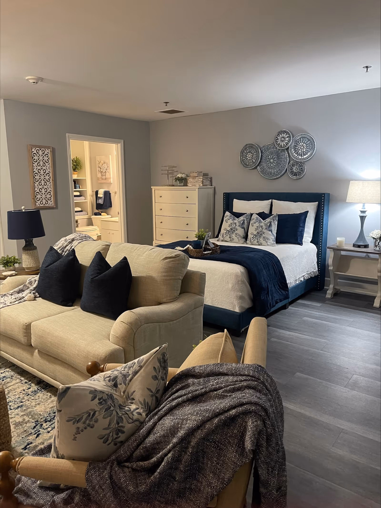 A cozy and well-decorated senior living bedroom with a blue upholstered bed, white and blue bedding, and decorative pillows. A beige sofa with dark blue pillows faces the bed. There is a wooden dresser against the wall and a bedside table with a lamp. The room has gray walls and wood-look flooring. An open doorway leads to a bathroom with towels and decor visible.