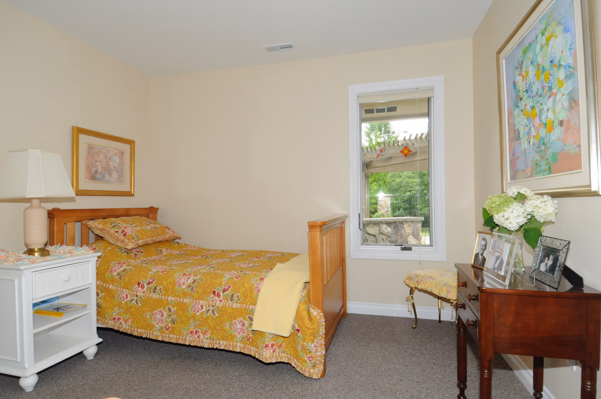 A small bedroom with a wooden single bed dressed in a yellow floral bedspread, a white nightstand with a lamp, a window, and a dresser topped with flowers and framed photos.