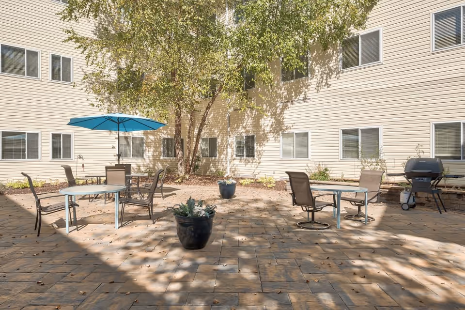 Sunlit courtyard patio with tables, chairs, a blue umbrella, potted plants, and a grill in front of a beige multi-window building.