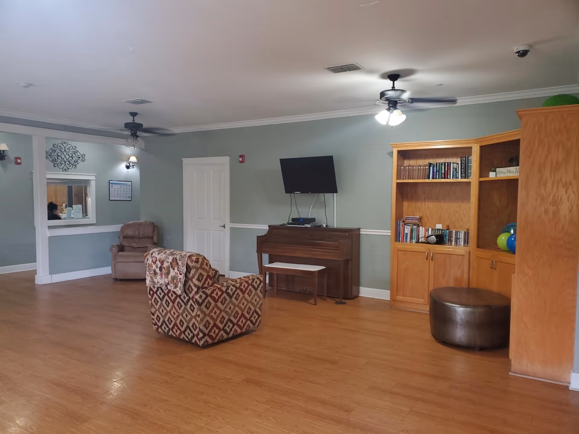 A spacious living room area with light green walls and wooden flooring. The room features a patterned armchair with a blanket draped over it, a beige recliner chair, a wooden piano with a bench, a wall-mounted TV above the piano, and a large wooden bookshelf filled with books and decorative items. Ceiling fans with lights are mounted on the ceiling, and there is a small window opening in the wall on the left side.