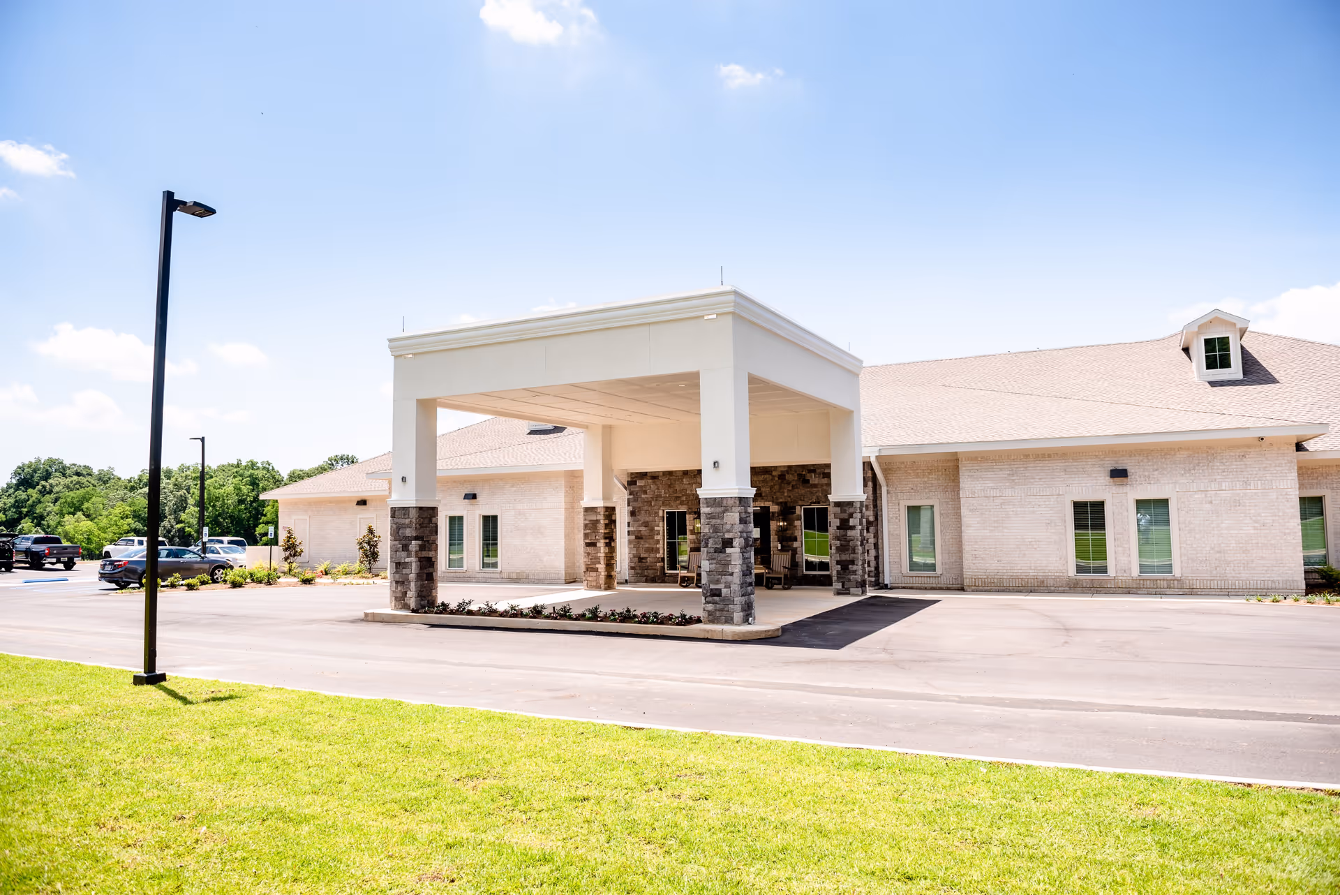 Exterior view of Carillon Oaks Mobile facility showing a covered entrance with stone pillars, a parking lot with several cars, green grass in the foreground, and a clear blue sky.