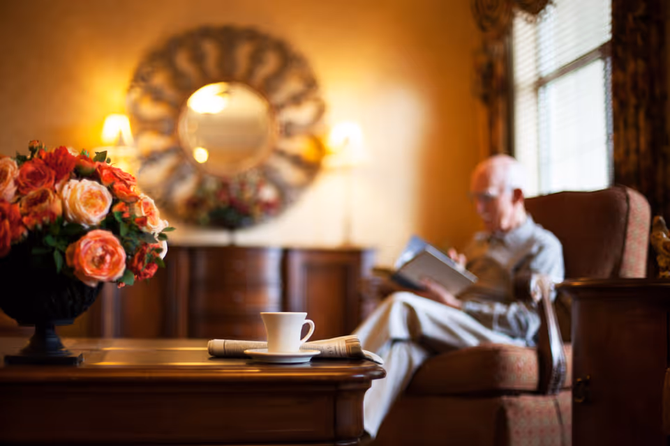 A cozy living room with a wooden coffee table in the foreground holding a cup and saucer and a folded newspaper. A vase with a bouquet of pink and orange roses is also on the table. In the background, an elderly man is sitting in an armchair reading a book. The room is warmly lit with two wall lamps and a decorative round mirror above a wooden cabinet.