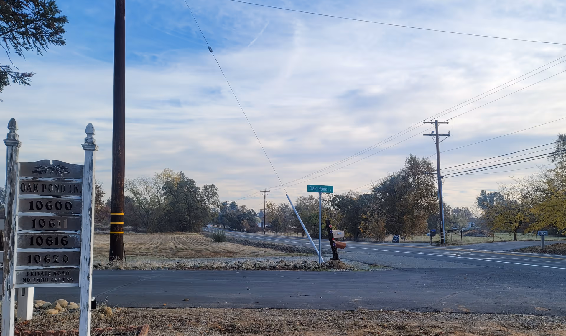 A rural road intersection with a wooden signpost on the left listing addresses on Oak Pond Lane. The sign reads 10600, 10611, 10616, and 10620. There are trees and open fields in the background under a partly cloudy sky. Utility poles and power lines run along the road.