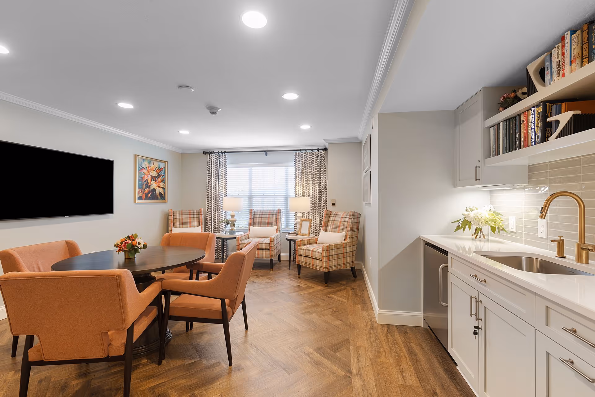 A cozy living room area with a round wooden table surrounded by four orange upholstered chairs. In the background, there are three plaid armchairs with white cushions near a window with patterned curtains and two table lamps. To the right, there is a small kitchenette with white cabinets, a sink with a gold faucet, a dishwasher, and shelves with books and decorative items. The floor has a wood pattern and the walls are light-colored.