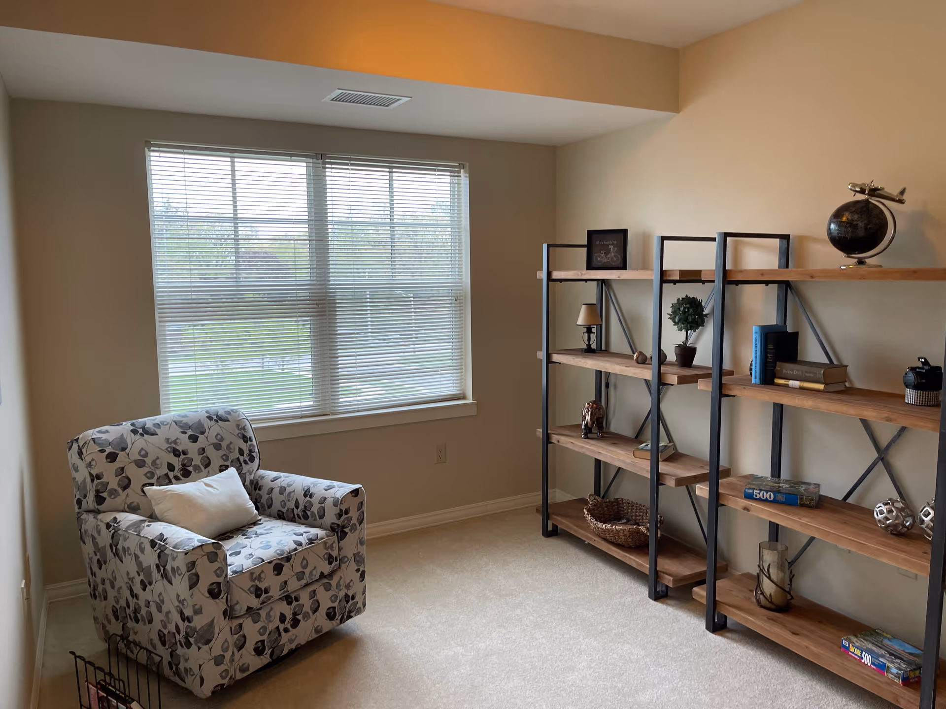 A cozy room with a large window covered by blinds, a patterned armchair with a white pillow, and two wooden shelves with black metal frames holding various decorative items including books, a globe, a small lamp, and puzzle boxes.
