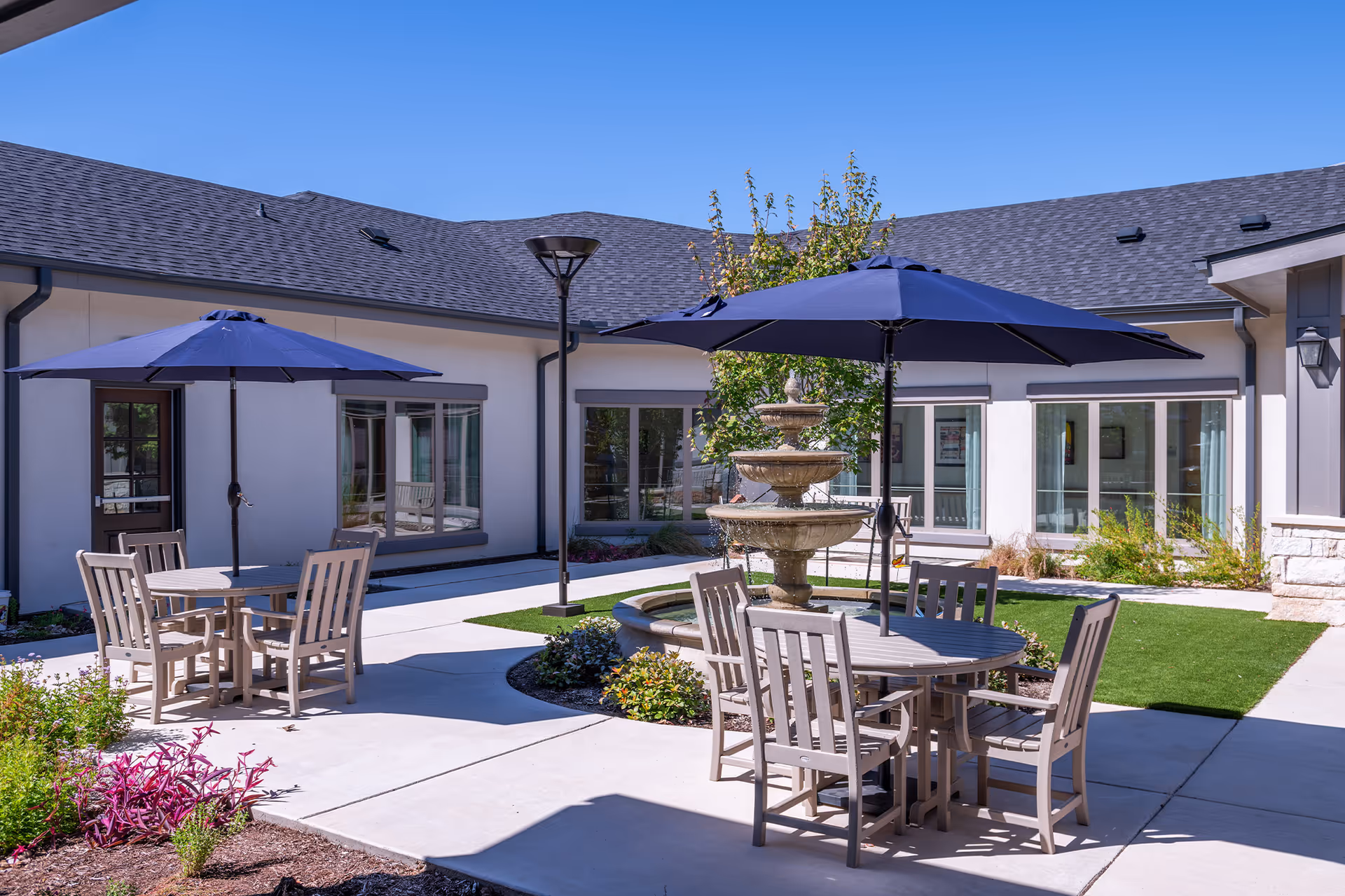 Sunny courtyard with round tables and blue umbrellas arranged around a central fountain in front of the senior living facility.