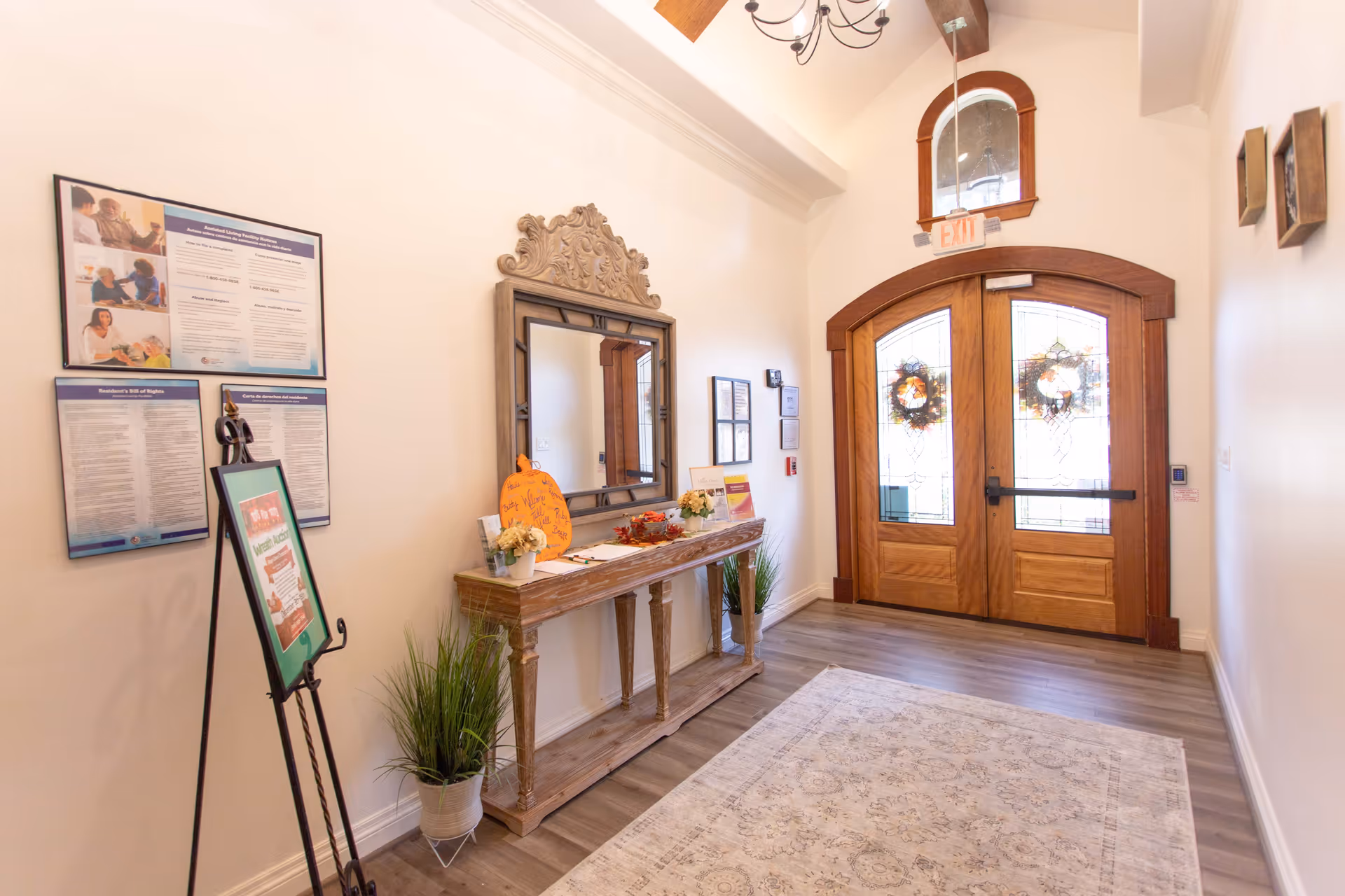 Entrance area of Village Green Memory Care Community Kingwood featuring double wooden doors with decorative glass and wreaths, a wooden console table with a large ornate mirror above it, potted plants on either side of the table, informational posters on the wall, and a light-colored patterned rug on the floor.