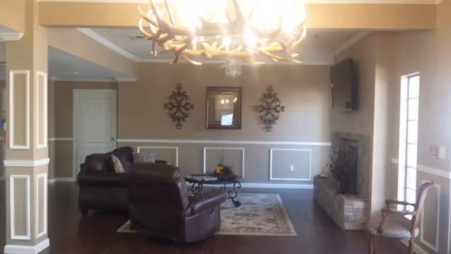 A cozy living room with beige walls and white trim, featuring a dark leather sofa and recliner, a decorative area rug, a coffee table with a floral centerpiece, a wall-mounted TV above a stone fireplace, and an antler chandelier hanging from the ceiling.