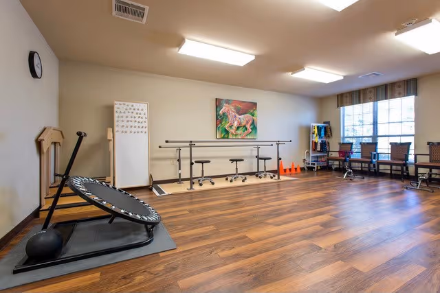 A spacious rehabilitation room with wooden flooring, exercise equipment including a mini trampoline and parallel bars, chairs lined up near a large window, and a colorful painting of horses on the wall.
