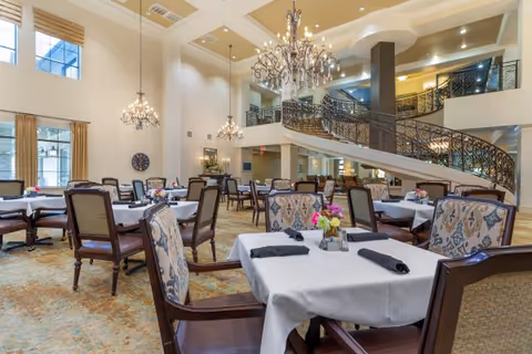 A spacious and elegant dining room with multiple tables covered in white tablecloths, each set with black napkins and small floral centerpieces. The room features high ceilings, large windows, chandeliers, and a decorative wrought-iron staircase leading to an upper level.