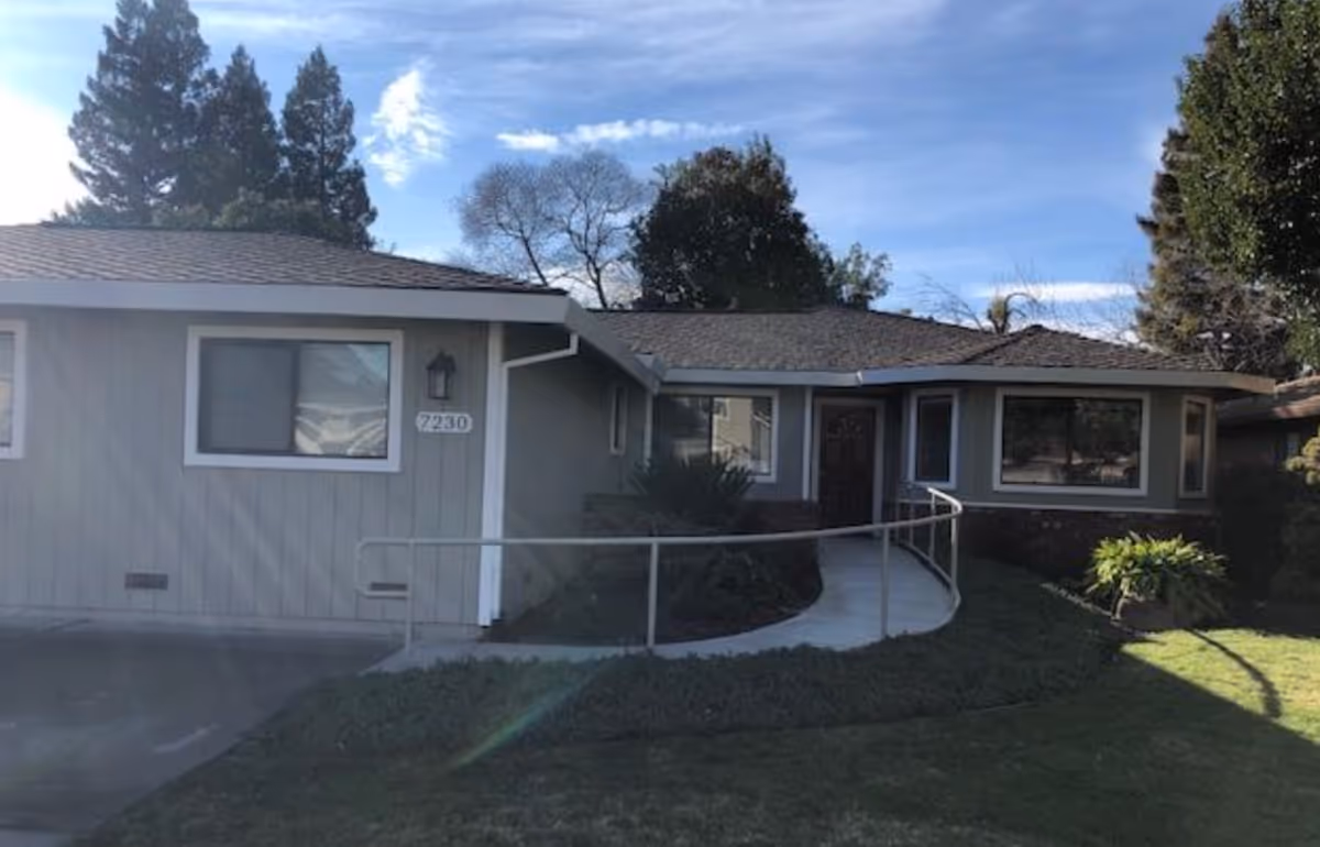 Single-story residential building with gray exterior walls, white trim around windows, a dark shingled roof, and a curved concrete ramp with metal railings leading to a wooden front door. The building is surrounded by grass, bushes, and trees under a partly cloudy blue sky.