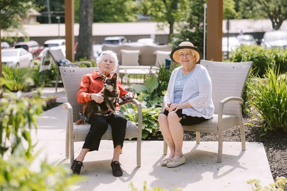 Two elderly women sitting outdoors on wicker chairs in a garden area. One woman is wearing a red jacket and holding a small dog, while the other woman is wearing a white cardigan and a sunhat. There are plants and greenery around them, with a parking lot and other outdoor furniture visible in the background.