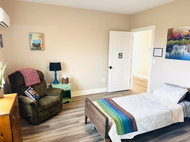 Sunlit bedroom with a single bed, recliner chair, nightstand and colorful throw and artwork.