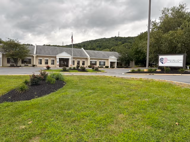 Exterior view of Highland Manor, a single-story building with a gray roof and beige walls, surrounded by a parking lot and landscaped greenery. An American flag is flying on a flagpole in front of the building, and a sign with the Highland Manor logo is visible on the right side near the entrance.