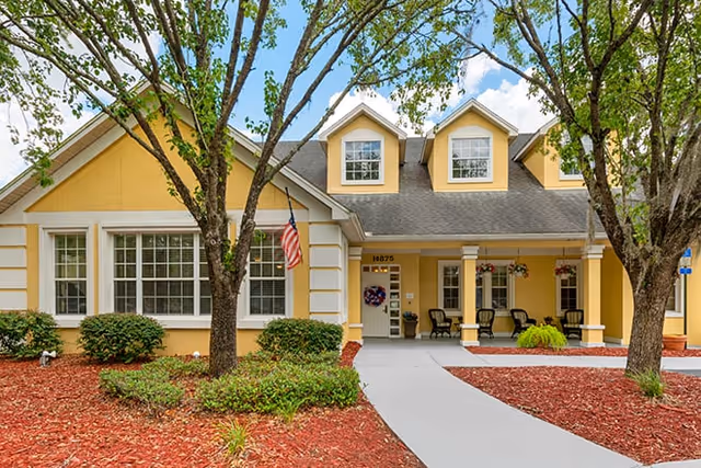 Front exterior of a yellow senior living building with a covered entrance, American flag, trees, and a paved walkway.