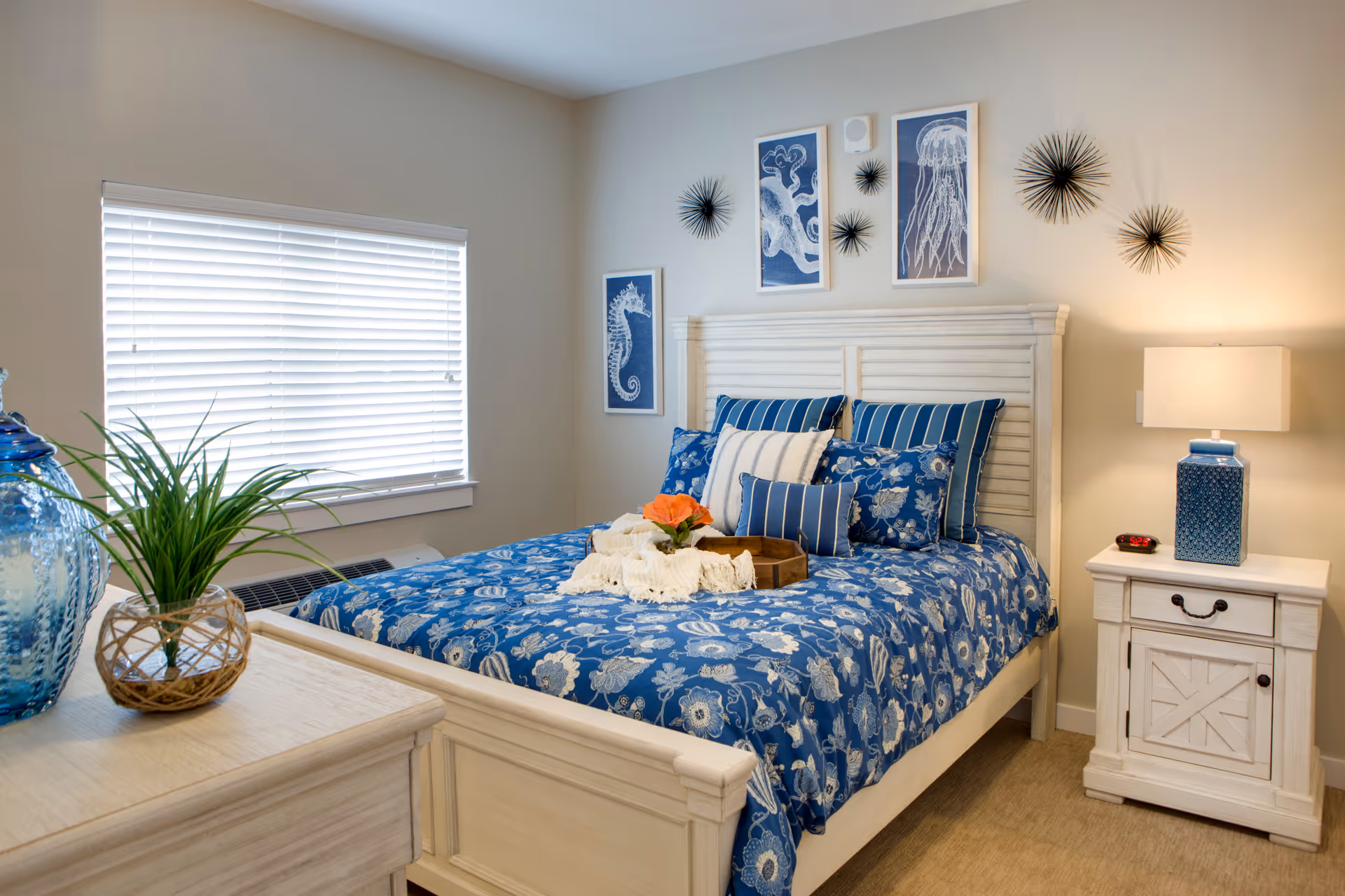 A cozy bedroom with a white wooden bed frame and blue floral bedding. The bed is decorated with multiple pillows in blue and white patterns. Above the bed are three framed sea creature artworks and several black decorative wall pieces. A white nightstand with a blue patterned lamp and a digital alarm clock sits to the right of the bed. A window with white blinds is on the left wall, and a dresser with a blue glass vase and a small plant is partially visible in the foreground.