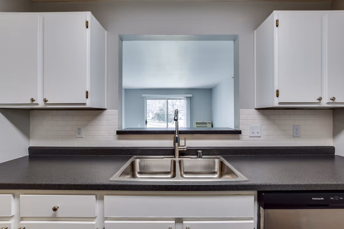 View of a kitchen countertop with a double stainless steel sink and a modern faucet. White cabinets with silver knobs are above and below the countertop. A pass-through window looks into a living room area with a large window and sliding glass door letting in natural light.
