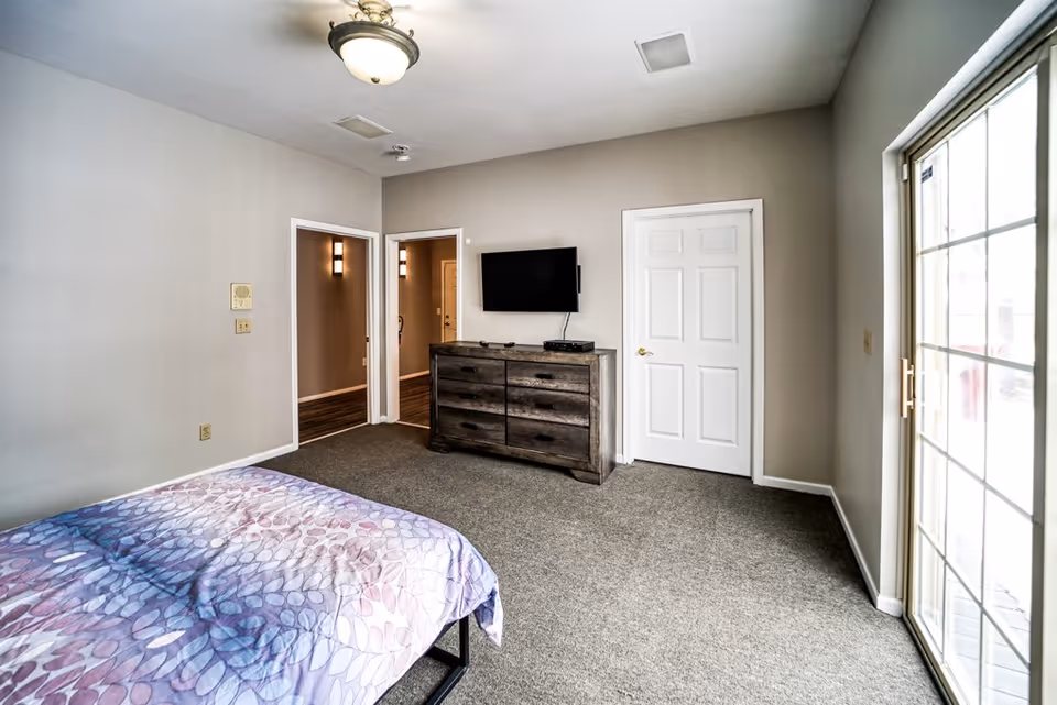 A bedroom with a bed covered in a purple patterned bedspread, a wooden dresser with a flat-screen TV mounted above it, two doors, and a large windowed sliding door letting in natural light.