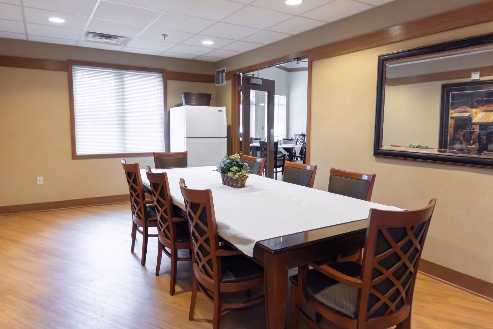 A dining room with a long wooden table covered with a white tablecloth and surrounded by eight wooden chairs with cushioned seats. A small flower arrangement is placed in the center of the table. There is a large window with blinds on the left wall and a white refrigerator near the window. A large mirror hangs on the right wall reflecting part of the room. The floor is wooden, and the ceiling has recessed lighting. An open doorway leads to another room with more tables and chairs.