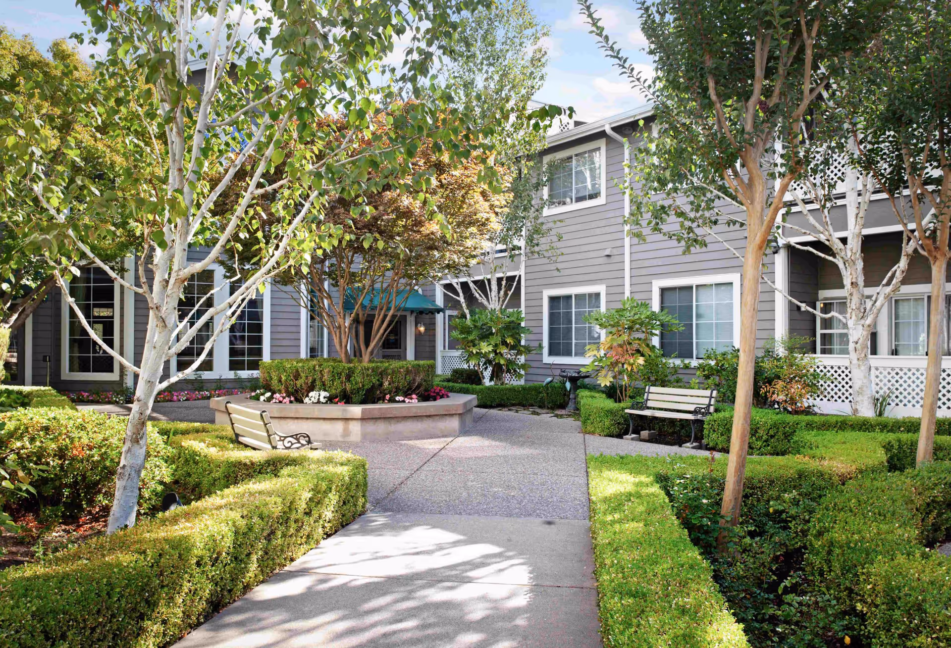 A landscaped outdoor courtyard area at Ivy Park at Oakland Hills featuring trimmed hedges, several trees, flower beds, two benches, and a paved walkway leading to a gray two-story building with white-trimmed windows.
