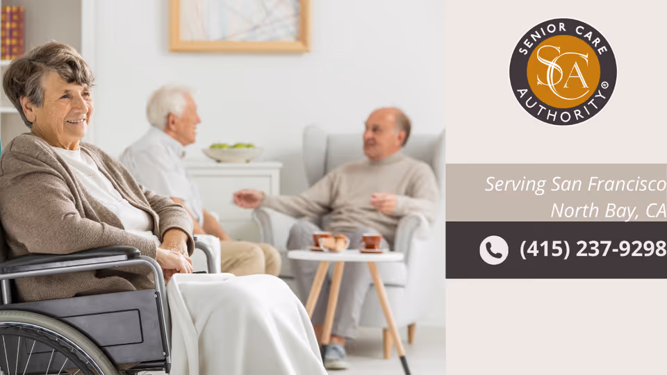 An elderly woman sitting in a wheelchair smiling in the foreground, with two elderly men sitting and conversing in armchairs in a bright, cozy living room setting.