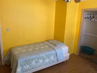 A small bedroom with yellow walls featuring a single bed with a green floral bedspread. There is an open closet on the right side with a few hangers and a green laundry basket on the floor.