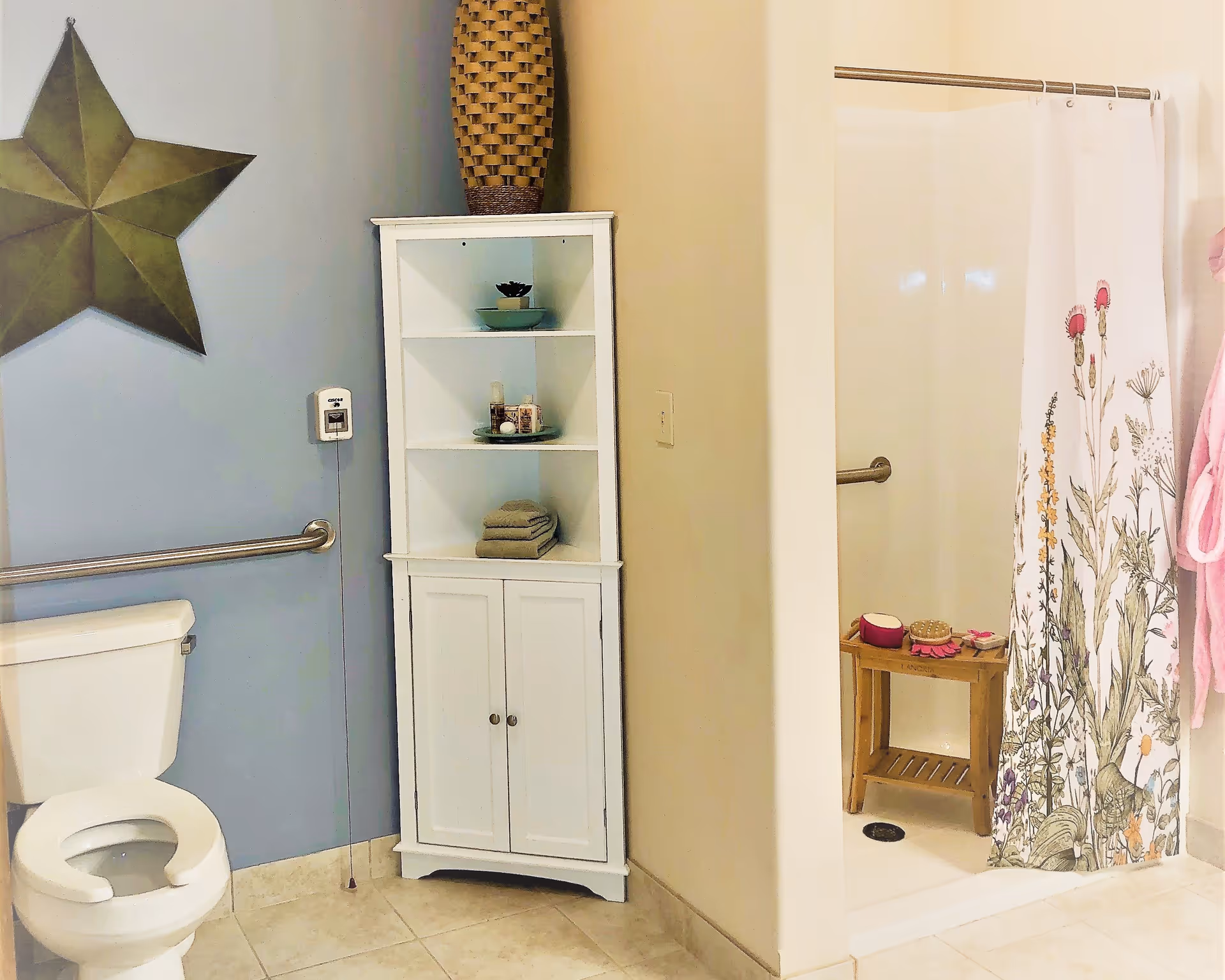 A bathroom with a white toilet, a white shelving unit holding towels and decorative items, a walk-in shower with a floral shower curtain, and a large decorative star on a blue wall.