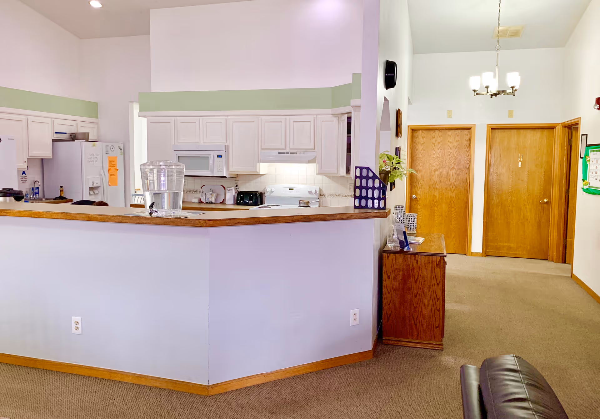 Open communal kitchen and seating area with a long countertop, appliances, and two wooden doors down a hallway.