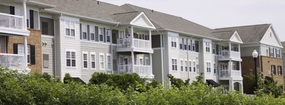 Exterior view of a multi-story residential building with white siding, brick accents, balconies, and numerous windows, surrounded by green bushes and trees under a clear sky.