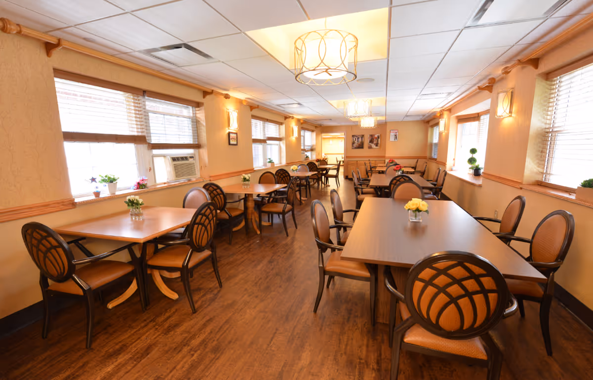 A well-lit dining room with multiple wooden tables and cushioned chairs arranged neatly. The room has large windows with blinds, small flower vases on each table, warm wall sconces, and ceiling lights providing a cozy atmosphere.