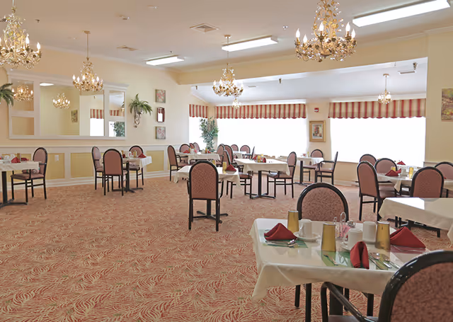 A spacious dining room with multiple tables covered in white tablecloths, each set with red napkins, cups, and utensils. The room features patterned carpet, several chandeliers hanging from the ceiling, large windows with red and white striped valances, and framed artwork on the walls.