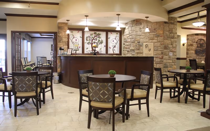 A cozy dining area in a senior living facility featuring round tables with cushioned chairs arranged neatly. The room has a warm ambiance with stone accent walls, pendant lights hanging from the ceiling, and decorative stained glass windows behind a wooden counter. Small green plants are placed on each table, and there is a seating area visible through an open doorway.