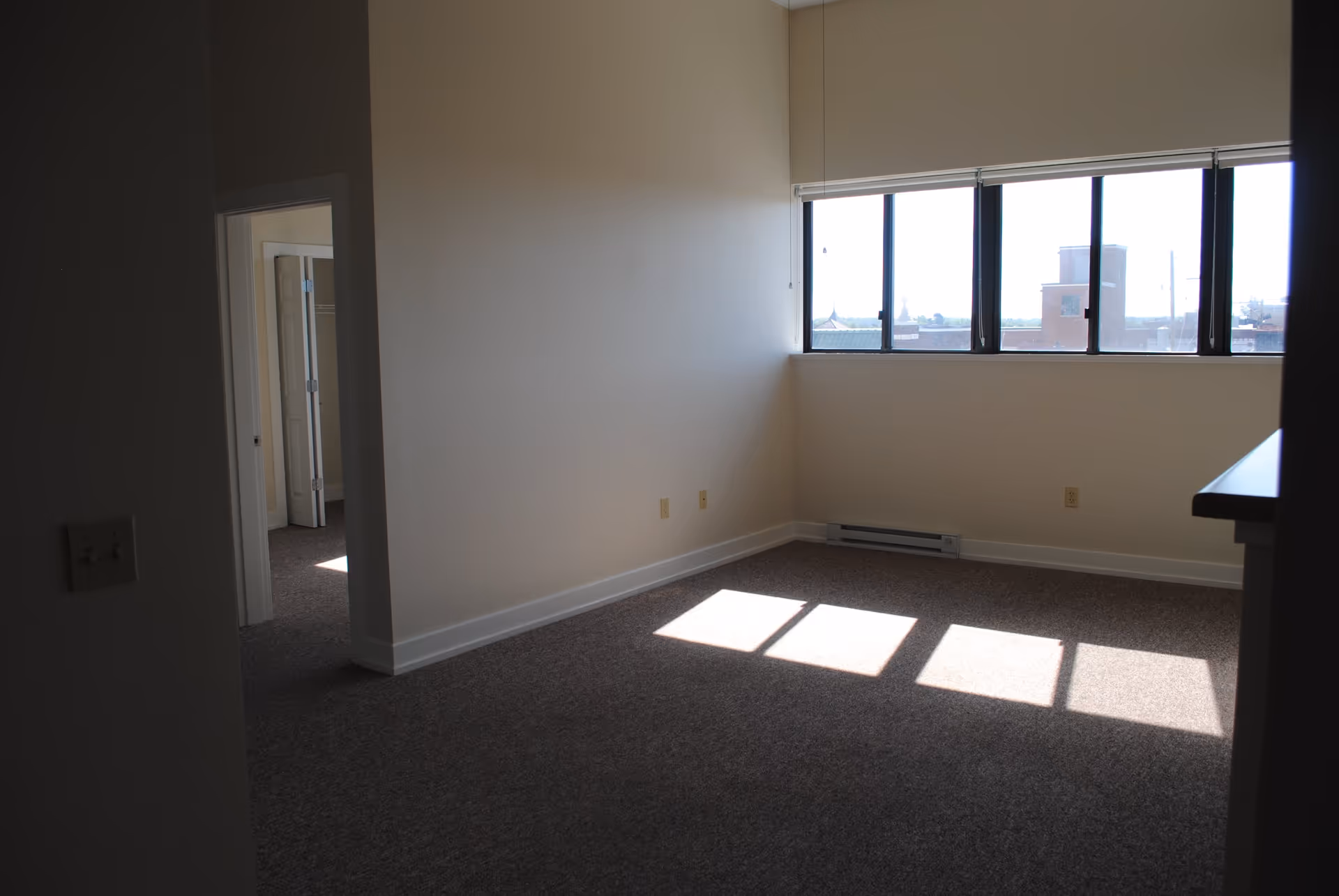 Empty room with beige walls and carpeted floor, large windows letting in sunlight, and an open doorway leading to another room with a closet.