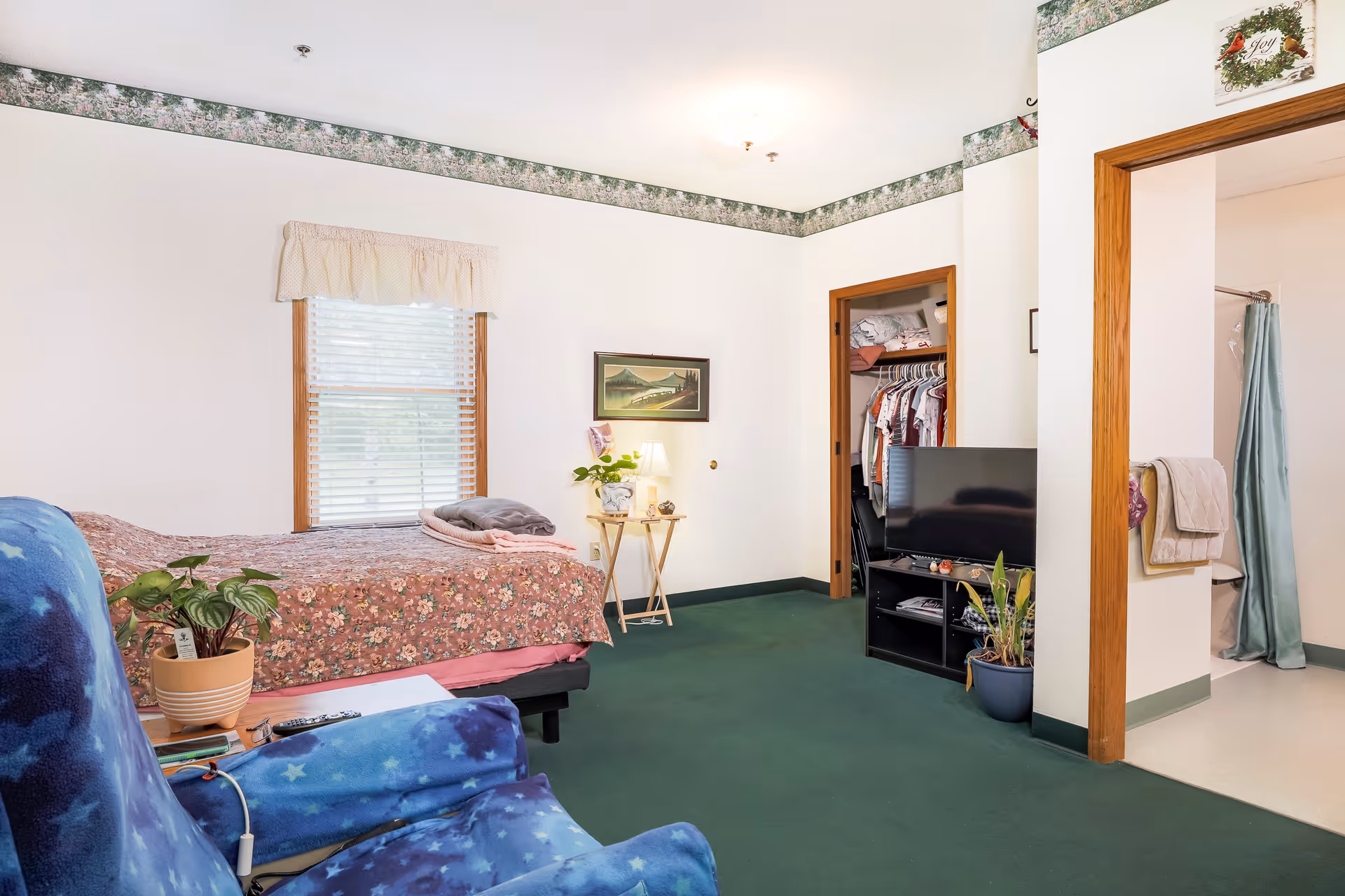 A senior living bedroom with a floral bedspread on the bed, a blue armchair with star patterns, a small wooden side table with a lamp and plant, a window with blinds and a valance, a closet with hanging clothes, a TV on a black stand, and an open doorway leading to a bathroom with a shower curtain and towels.