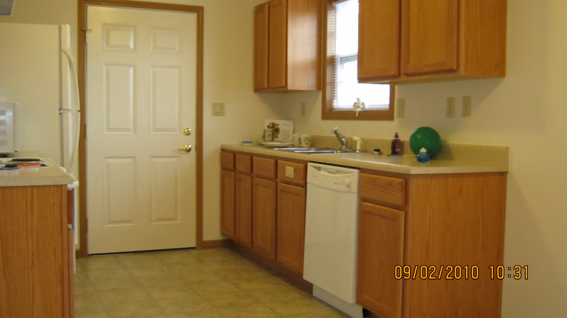 Interior view of a kitchen with wooden cabinets, a white dishwasher, a white refrigerator, a microwave, a sink under a window, and a door at the end of the room. The floor is tiled and the walls are light-colored.