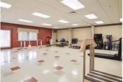 A spacious rehabilitation room with exercise equipment including parallel bars, a treadmill, therapy balls, and a stationary bike. The room has a tiled floor with red and white squares, beige walls, and a large window with white curtains allowing natural light to enter.