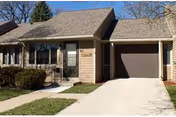 Single-story townhouse-style building with a front door, attached garage, driveway, and small front yard with shrubs.