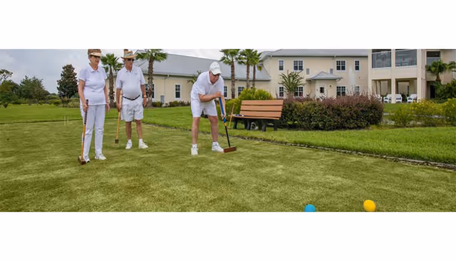 Three people playing croquet on a grassy lawn in front of a multi-story residential building with a bench and palm trees.