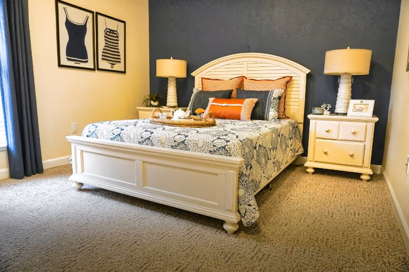 A cozy bedroom with a white wooden bed frame and a floral patterned bedspread. The bed is adorned with multiple pillows in shades of orange and gray. There are two matching white nightstands on either side of the bed, each with a lamp. The wall behind the bed is painted dark blue, while the other walls are light-colored. Two framed pictures of swimsuits hang on the left wall, and a window with dark curtains is partially visible.