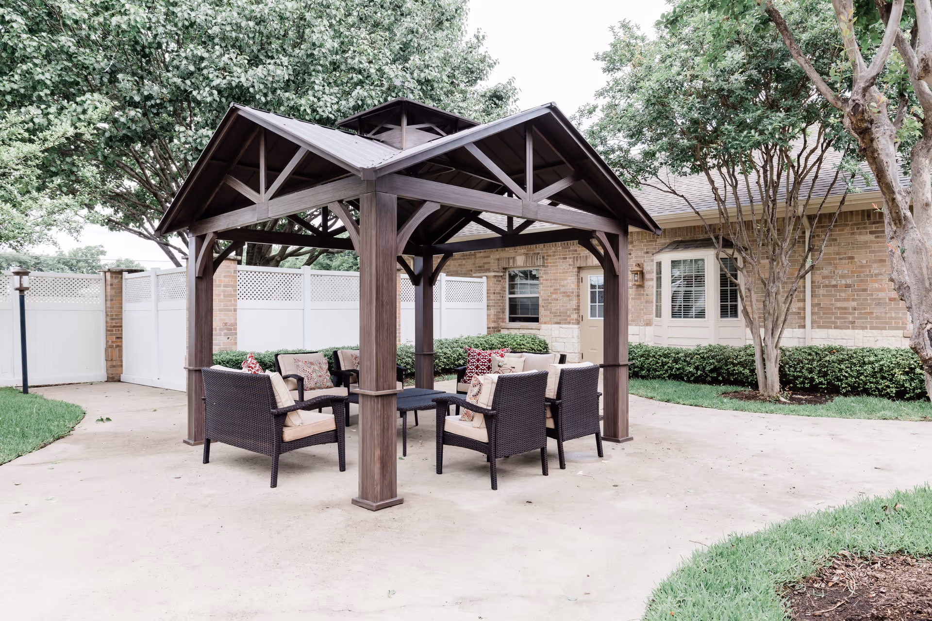Outdoor seating area with a wooden gazebo structure covering cushioned wicker chairs and a small table, surrounded by a concrete patio, green grass, trees, and a brick building with windows and a door in the background.