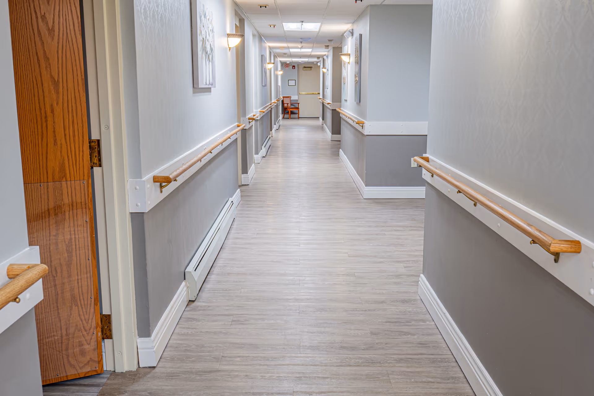 A long, clean hallway in a senior living facility with light gray walls, wooden handrails on both sides, light wood flooring, wall-mounted lights, and a wooden door partially open on the left side.