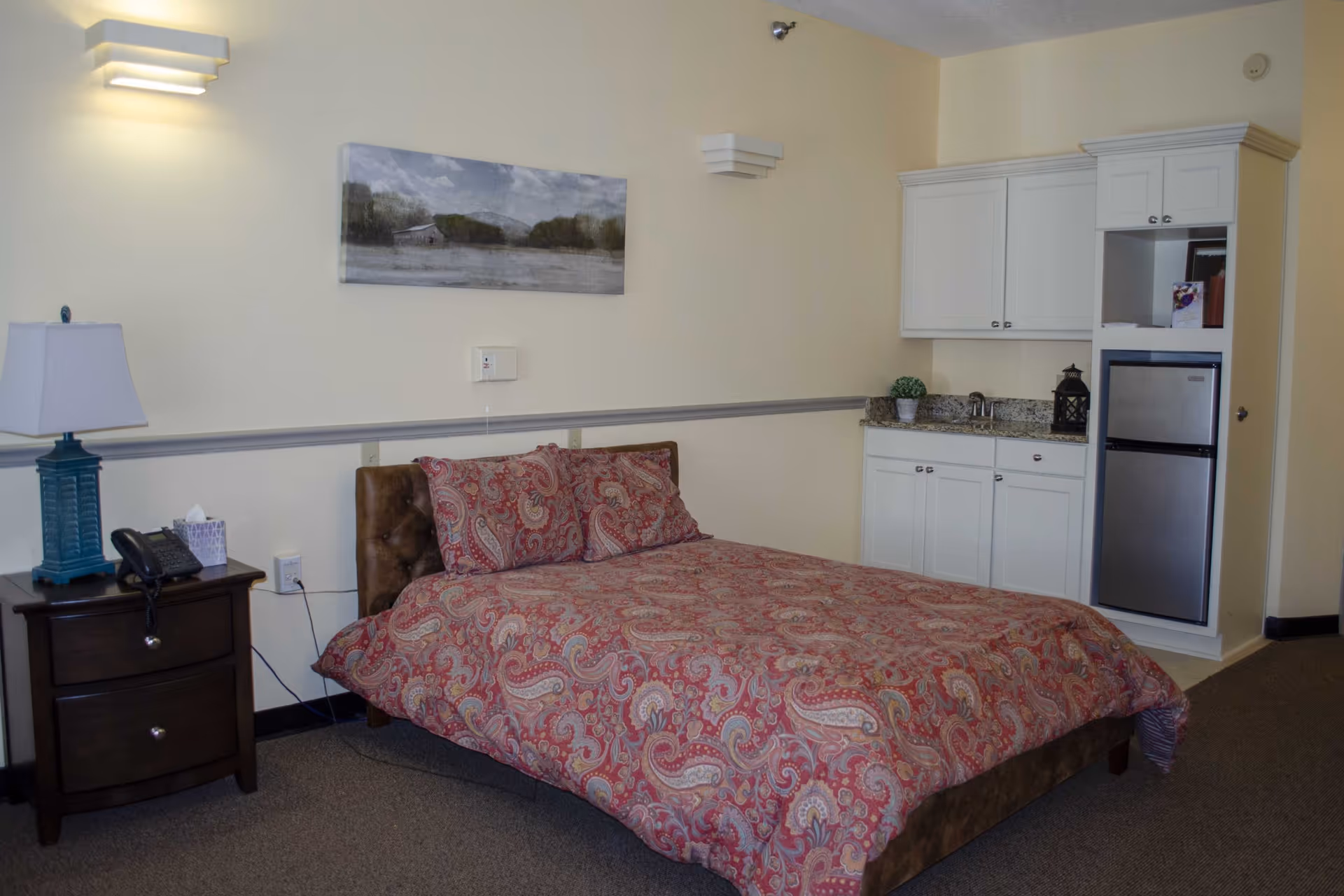 A furnished assisted living studio bedroom with a bed, nightstand, lamp and a small kitchenette with cabinets and a mini-fridge.