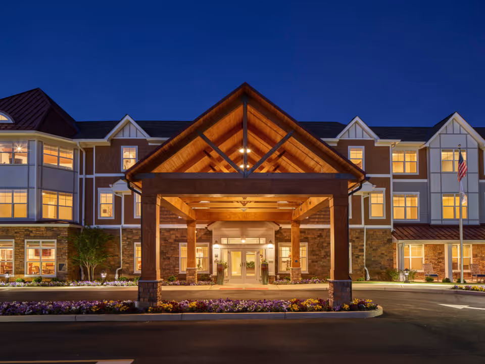 Illuminated front entrance of a senior living building at dusk with a covered porte-cochère and flowerbeds.