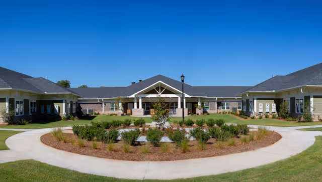 Front entrance of a single-story senior living facility with a circular landscaped courtyard under a clear blue sky.