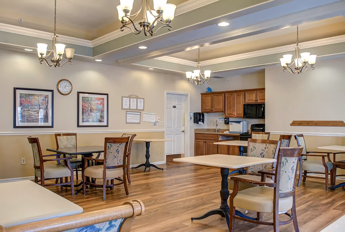 A well-lit dining room with several tables and chairs arranged neatly. The room features wooden flooring, beige walls with white trim, and three chandeliers hanging from the ceiling. On one wall, there are two framed landscape paintings and a clock. In the background, there is a kitchenette area with wooden cabinets, a microwave, and a sink.